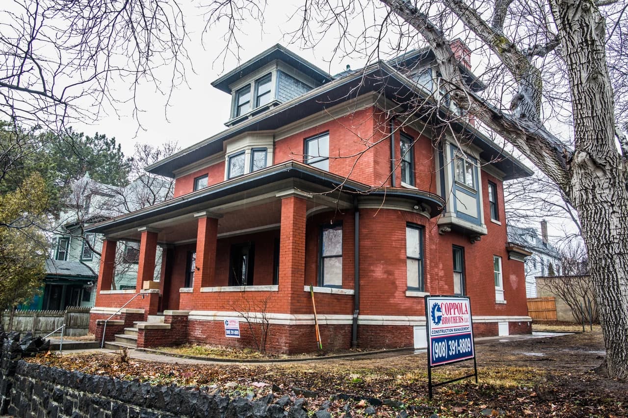 Red brick house with a deep front porch, projecting bay windows, and a dormer beneath leafless trees.
