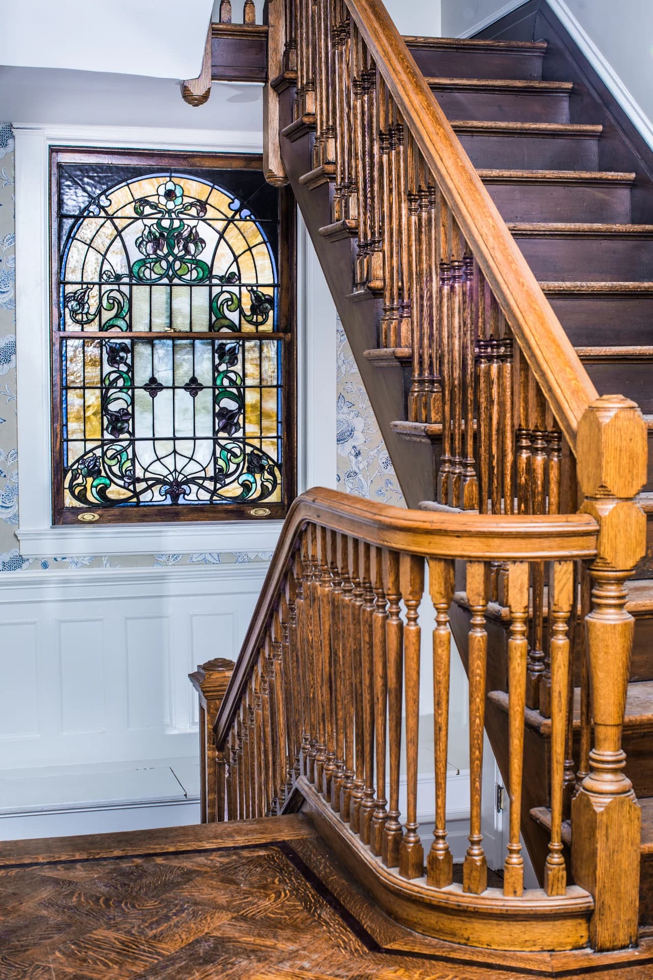 Curved wood stair railing and turned balusters beside a tall decorative stained-glass window.