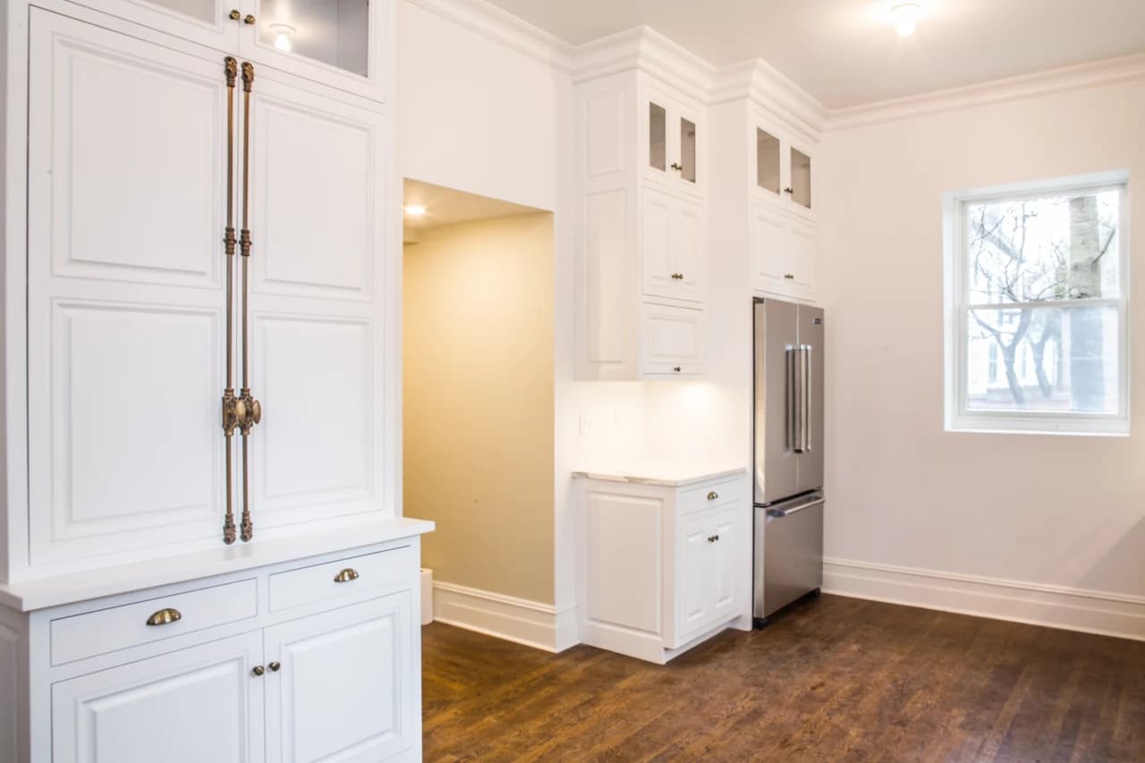 Tall white kitchen cabinets with decorative metal hardware beside a stainless-steel refrigerator.
