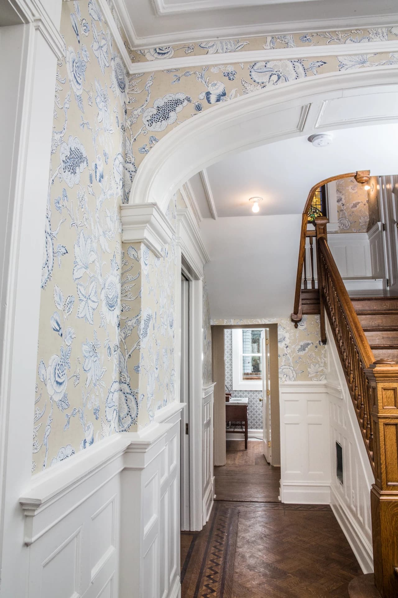 Arched hallway with floral wallpaper, white paneled wainscoting, and patterned wood flooring beside a staircase.