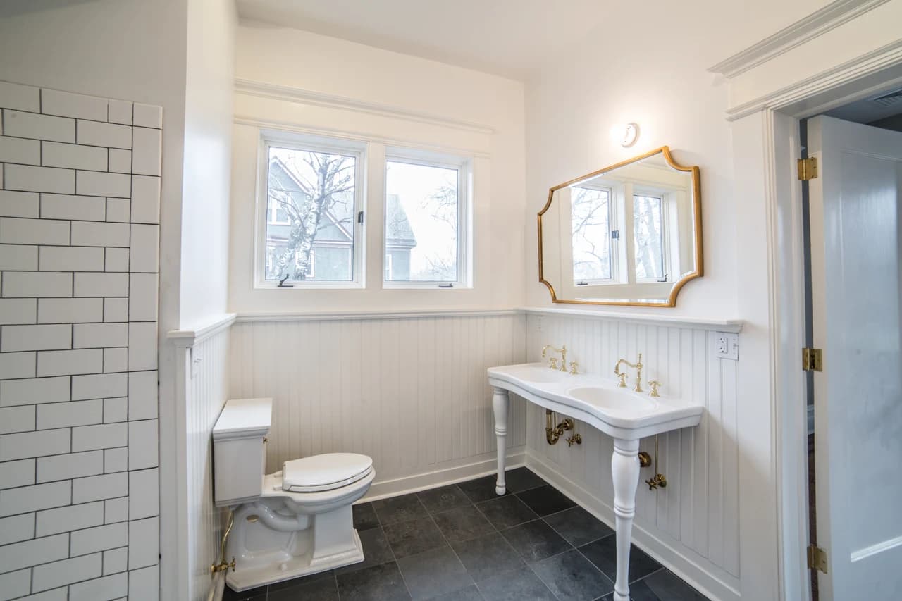 White bathroom with a double console sink, brass fixtures, a gold-framed mirror, and dark floor tile.
