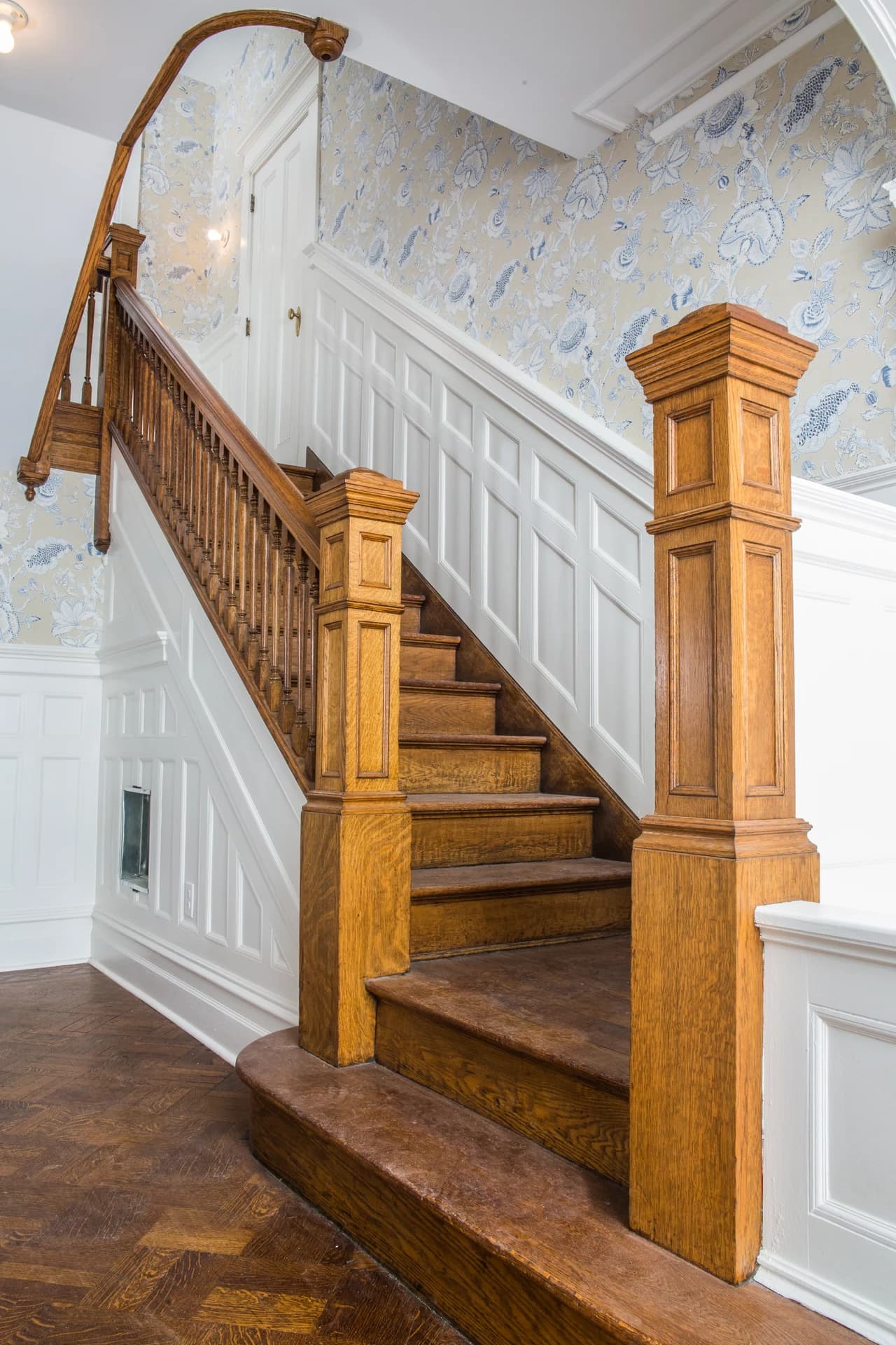 Wood staircase with paneled newel posts, a curved lower step, and floral wallpaper above white wainscoting.